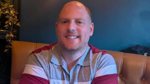 Colin Jones Colin. He is bald and wears a striped polo shirt as he sits on a brown leather chair.