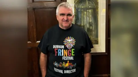 Handout Stephen Cronin standing in front of a picture, wearing a black Durham Fringe T-shirt. He has short grey hair.
