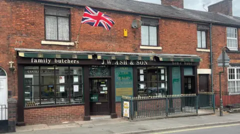 The outside view of a traditional butchers. A Union Jack flag hangs above the shop attached to terraced houses. The signage across the top of the shop reads ‘FAMILY BUTCHERS: J.W. ASH & SON’