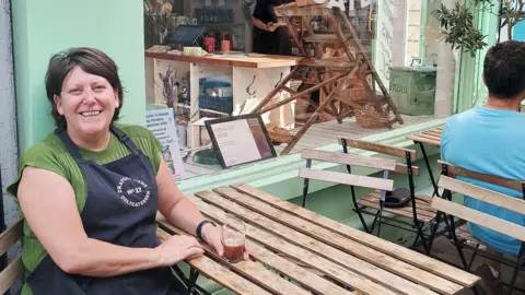 Samantha Morgan sits outside her business and holds a drink with her left hand. She has short brown hair and is wearing a green top with a black apron over the top.