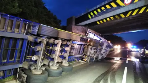 Leicestershire Police A large lorry is on its side after hitting the A5 bridge in Hinckley, because it's too big to get through. It's dark and you can see vehicle lights in the distance as other drivers can't get through due to the accident. 