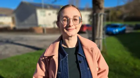 A young woman with big round glasses and brown hair tied back. She is wearing a pale pink jacket and standing on a housing estate.