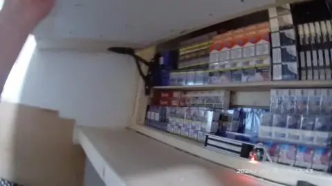 A cupboard full of illegal tobacco products and vapes. The still, taken from a police body camera, shows a hidden compartment being lifted open and packets of cigarettes stacked on a shelf.