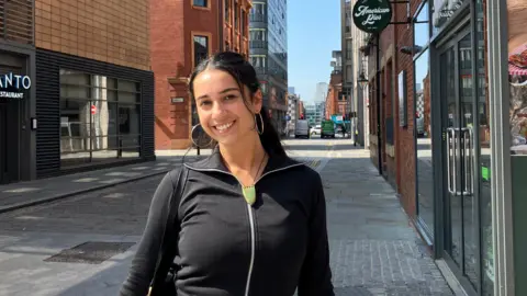 BBC/@sufia.grace Sufia in the foreground wearing a black zip-up top and light skirt, carrying a black shoulder bag, standing on a paved side street. Behind, a narrow roadway leads to an intersection with traffic lights and a green van. Red-brick and modern buildings line both sides, including a glass-fronted shop with the sign “American Pies”. A large blue-painted utility structure sits to the left. The street has kerbs, paving slabs, and no visible bins, with clear road markings and a few pedestrians in the distance under a bright sky.