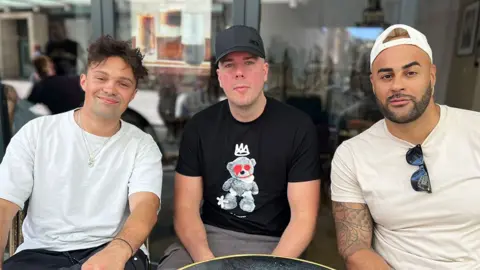 BVNQUET (left) wears a white t-shirt and sits next to Neutrino (right) of Solid Crew, who wears a white t-shirt, and Oxide (middle), who wears a black t-shirt.