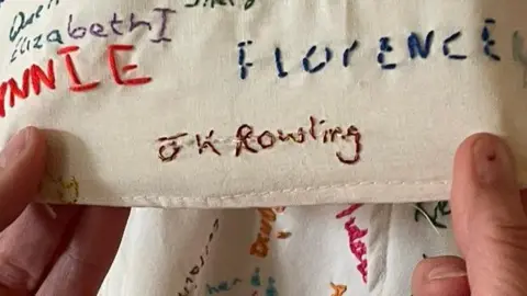 A pair of hands are holding a piece of fabric covered in embroided names. The one being held prominently to the camera is "JK Rowling"