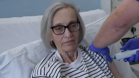 Chris Jones receives a vaccine into her left arm. She is lying in a hospital bed and wears a white and black striped top. She has grey hair and wears dark-framed glasses.