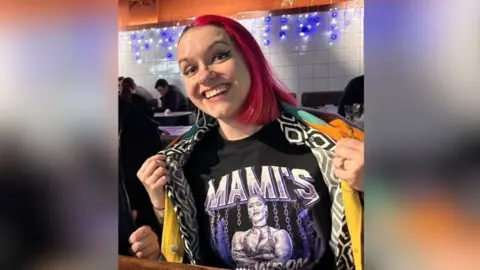 Bethany Crooks Person with long pink hair seated indoors at a hospitality venue at a wooden table, wearing a black T-shirt with a graphic design featuring a muscular womanin chains and the text ‘MAMI’S’ in large purple letters. The person is pulling open a patterned jacket to reveal the shirt. In the background, there are white tiled walls, blue decorative lights, and other people seated at tables.