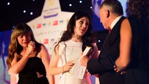 Dougie Allward/Made for impact Aliese has long dark hair and is wearing a white dress as she stands on stage with her prize. She is talking to a man on stage.