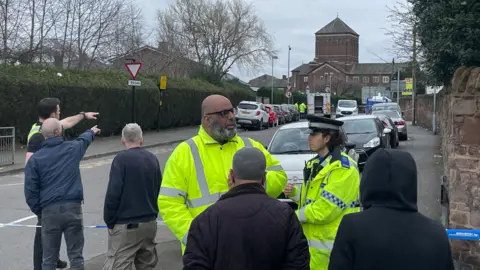 A group of people, some wearing hi-vis jackets and police uniform, stand behind a police cordon on a residential road.