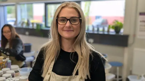 A woman stands in a craft studio. She has long straight blonde hair and glasses, and she is wearing a black top and a beige apron. A woman is blurred in the background sat near a table with pots and paintbrushes.