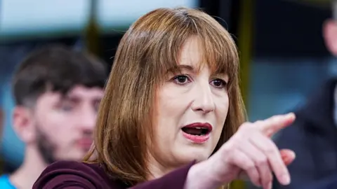 Rachel Reeves with her arm outstretched, pointing. She is wearing a purple top, with people standing behind her out of focus. 