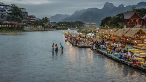 Getty Images People drink and enjoy food on a riverbank in the town of Vang Vieng in Laos. There as some people swimming in the river and a string of lights highlights dozens of people sat on tables.
