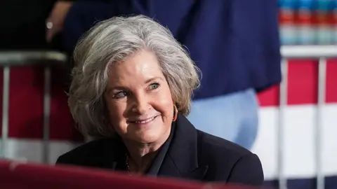 Susie Wiles, sporting a silver bob and a black coat, smiles.