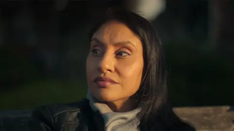 Netflix still from the Netflix documentary featuring Kirat, a woman sitting outside and looking to the left. She is wearing a black leather jacket and a gray scarf.