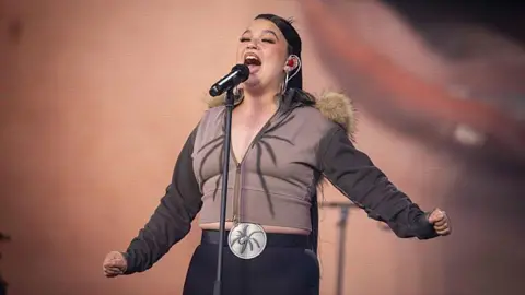 Lola Young is singing into a microphone on stage. She wears a brown zip up jacket with a fur collar hood paired with black trousers.