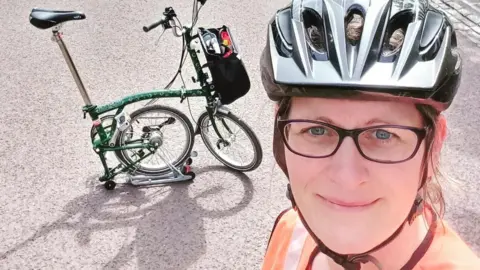 LCC Cyclist Louise Whittle in a selfie. She is wearing glasses, a cycling helmet and an orange hi-vis top. Her green bike, one of those ones that folds up, is on the road behind her.