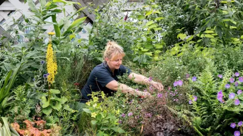 A female gardener, wearing a navy blue polo shirt, crouches down as she tends to colourful shrubs and flowers at the Castlefield Viaduct urban park in Manchester.