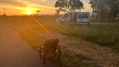 Billy Whiston A wheelchair on a road looking out to the horizon where the sun is setting. Billy's van is parked in the background.