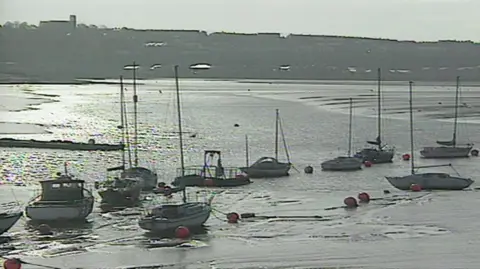 The image shows several boats moored in the mudflats of Cardiff Bay before the barrage was built. The Penarth headland can be seen in the background.