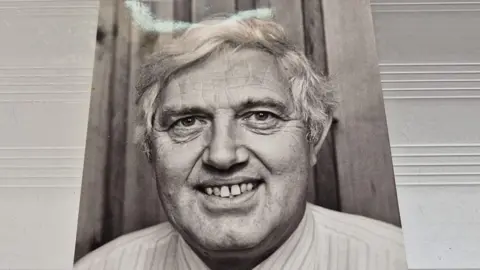 James Baillie A black and white photo cut out showing a man with white hair, smiling into the camera.