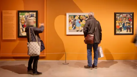David Levene/Art Fund 2026 The photo shows people looking around the Beryl Cook: Pride and Joy exhibition, which ran between January and May. There is a bright orange wall on which three pieces of Beryl Cook art are hung. One woman is taking photos.