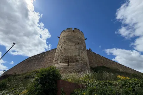 A view looking up towards the top of an imposing, round castle tower, with high walls and battlements to either side. The castle is built of stone on a raised green bank, with wild flowers growing in the foreground. The sky is blue with white clouds.