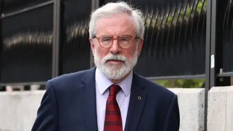 Gerry Adams has white hair and a beard. He wears brown glasses, a navy suit, a light shirt, a red tie, and a pin on his lapel