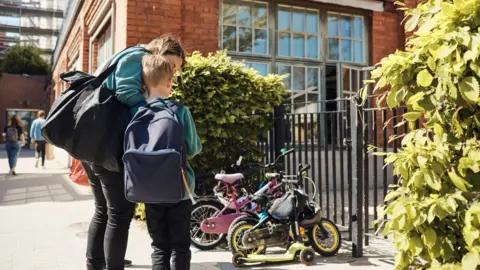 Getty Stock image of a mum comforting a child outside a school. The boy has short blonde hair and a large blue backpack. The mum is wearing jeans and a teal jumper, and carrying a black shoulder bag. There are bikes and scooters outside the red-bricked school, and a black gate.