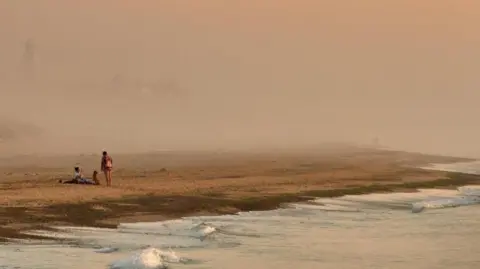 A photograph of Southwold beach showing people on a misty, windswept stretch of sand with the town's lighthouse just visible in the background


