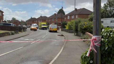 A residential street with police tape across the front. In the distance you can see several vehicles including an ambulance and a police van.