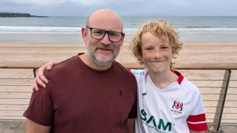Father and son Nigel (left) and Leighton Christie (Right). Nigel has a beard and is wearing a red top and black-rimmed glasses. Leighton is wearing an Ulster Rugby jersey and has long wavy blonde hair.