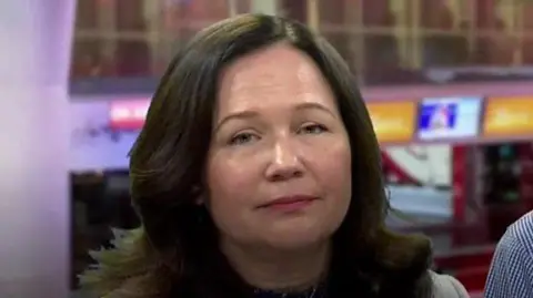 Tanya Ednan-Laperouse, with long dark hair, looking at the camera in a television news studio. A monitor is visible to the right.