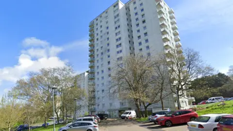 Google A tall apartment under clear blue skies. There is a small car park with a handful of cars parked in it. 