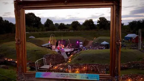 The Roman Theatre at Verulamium as seen through a picture frame as the sun goes down during a production. It shows a backdrop of distant large trees, and the stage nestles within the flint walls of the Roman remains