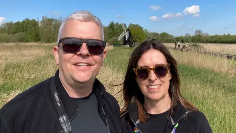 Dave Webster/BBC Ellis Selway with a woman colleague at Wicken Fen Nature Reserve with an Edwardian wind pump behind