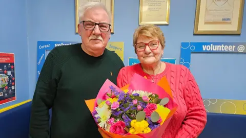 Graham Gedney in dark shirt and glasses stands next to his wife, Linda Gedney, who is wearing a pink jumper and has red glasses and is holding a bouquet of flowers