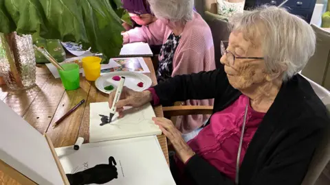 Cathy Killick/BBC An elderly woman with short grey hair and glasses is seated at a wooden table as she draws a black cat on a canvas. She wears a bright pink top under a black cardigan.