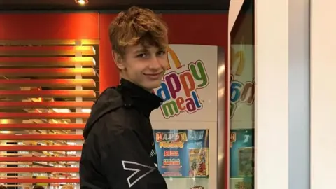 Jacob Crawshaw looking sideways at the camera while standing in front of a fast-food restaurant's self-service ordering board.
