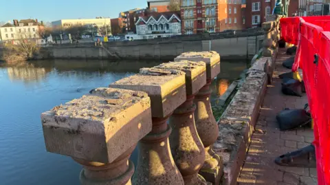 A damaged bridge with red barriers up. A river can be seen and buildings on the far side lining a road.
