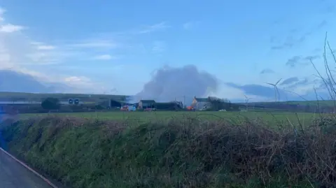 St Dennis Community Fire Station A view fop the farm from distance. A mass of smoke is billowing into the sky. There are two fire engines and three emergency vehicles visible next to the cluster of farm buildings. There is a hedge and a road in the foreground and a field between the person taking the photo and the farm.