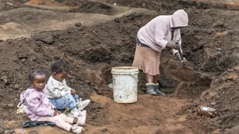 AFP/Getty Images A woman uses a spade to fill a bucket with soil next to her children as artisanal miners dig holes looking for gold in a patch of land outside Springs.