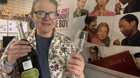 Phil Harrison/BBC Jo Holmes, manager of the Carlton Cinema in Westgate, stood in front of a poster for the latest Bridget Jones movie - Mad about the Boy. She is holding a champagne glass in her left hand and a bottle of prosecco in her right hand. 