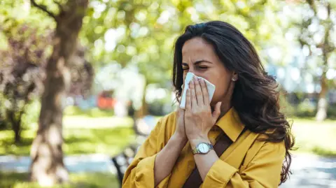 A woman with long brown hair, wearing a yellow shirt, holds a tissue to her nose as she sneezes, with trees in the background