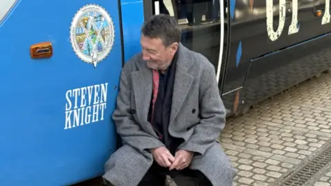 PA Media Steven Knight, wearing black suit and grey coat, is crouching by the side of a blue section of tram bearing his name. There is cobbled street beneath the tram and a drainage grid running alongside.
