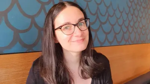 Name for Oxevision cameras to be banned in psychological well being sufferers' bedrooms Nell A woman sat down on a bench that is against a wall. She is smiling to camera and is wearing glasses. Behind her is blue wallpaper.