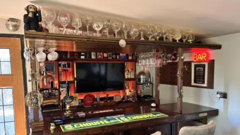 Steve Amos A small bar with runners and a beer pump on it. A number of glasses sit above the bar, and a TV is positioned behind it.