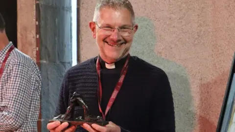 Dean John Witcombe wearing a black jumper smiling at the camera, stood outside in the evening. He is holding a small statue