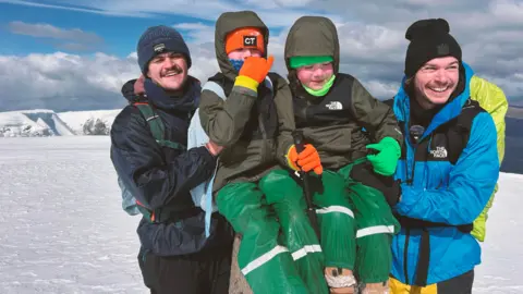 The Brothers Shed Two adults stood next to two young children on a mountain. They are all wearing hiking gear and look happy.