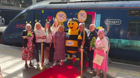 Isabella Norrison/BBC A group of people, many wearing colourful outfits and bee headbands, and someone dressed as a bee, standing in front of train on a railway platform.